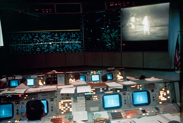 Date Created: 1969-07-20 Interior view of the Mission Operations Control Room (MOCR) in the Mission Control Center (MCC), Building 30, during the Apollo 11 lunar extravehicular activity (EVA). The television monitor shows astronauts Neil A. Armstrong and Edwin E. Aldrin Jr. on the surface of the moon.