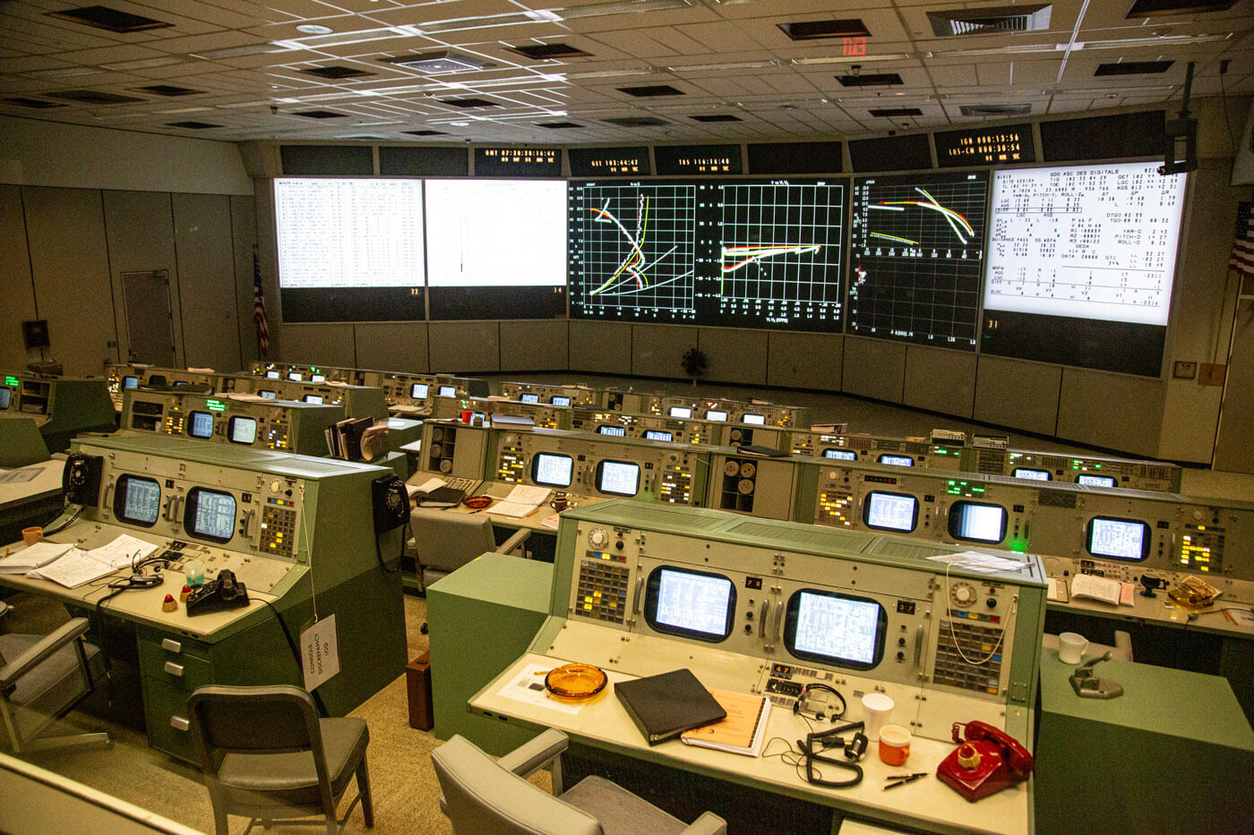 See the restored Apollo Mission Control Center with the actual flight control consoles brought back to life. The five large screens across the front of the room have been reactivated with projections to recreate the exact images seen during the Apollo 11 mission. See the console buttons illuminated and furnishings on the consoles including flight control manuals, ashtrays, pens, maps, coffee cups and headsets.