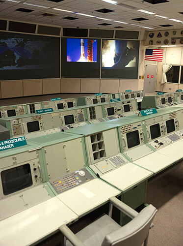 An overall view of the consoles used in Historic Mission Control. The consoles used by multiple flight directors contained no computing elements - they displayed only different information channels coming in from the mainframe. On the left side of the console is a pneumatic tube station through which controllers passed papers between consoles. Of the hundreds of buttons, monitors and controls located at these flight controller stations, many have gone missing or have become faded after decades of use.