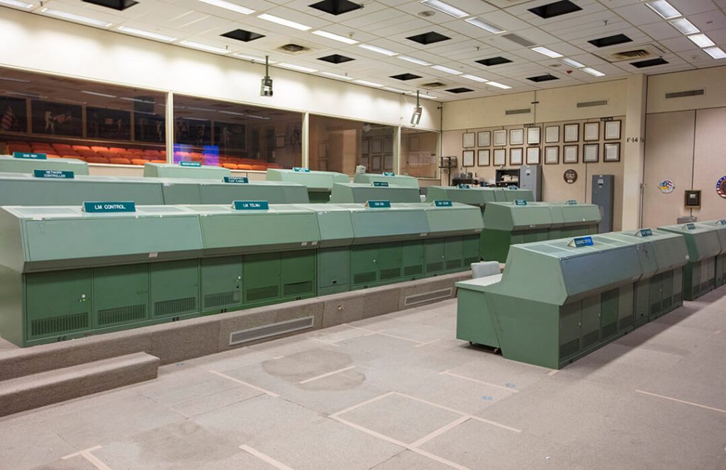 A front view of the iconic green consoles in Historic Mission Control. After decades of use, the carpet in the room has become stained, is deteriorating and is held together by tape.