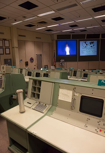 An overall view of the consoles used in Historic Mission Control. The consoles used by multiple flight directors contained no computing elements - they displayed only different information channels coming in from the mainframe. On the left side of the console is a pneumatic tube station through which controllers passed papers between consoles. Of the hundreds of buttons, monitors and controls located at these flight controller stations, many have gone missing or have become faded after decades of use.